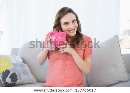 Smiling pregnant woman shaking pink piggy bank sitting on couch looking at camera