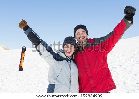 Portrait of a happy loving couple raising hands with ski board on snow in background against clear blue sky