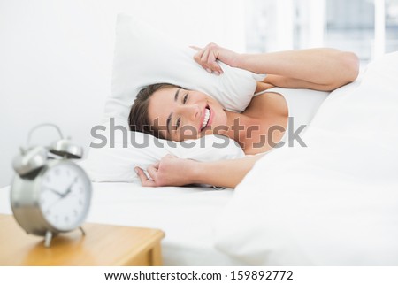Smiling young woman covering ears with pillow in bed and alarm clock on side table