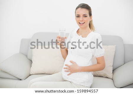 Peaceful pregnant woman holding a glass of water touching her belly smiling happily at camera