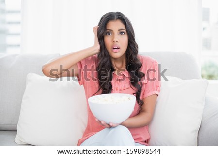 Shocked cute brunette sitting on couch holding popcorn bowl in bright living room