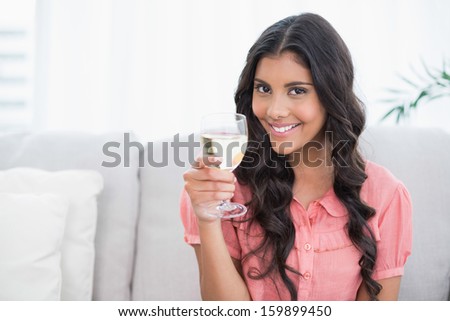 Smiling cute brunette sitting on couch holding white wine glass in bright living room