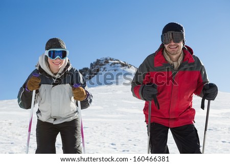 Portrait of a smiling couple with ski poles on snow covered landscape against clear blue sky
