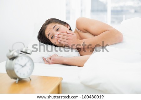 Shocked young woman in bed looking at alarm clock at home