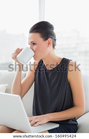 Beautiful well dressed young woman using laptop while drinking coffee on sofa at bright home