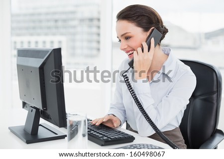 Smiling Elegant Businesswoman Using Landline Phone And Computer In A Bright Office