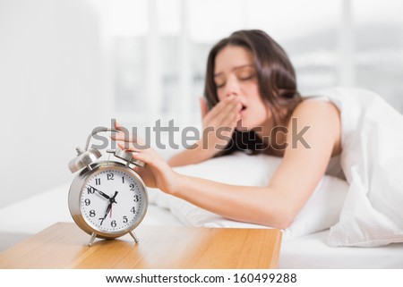 Young woman yawning while extending hand to alarm clock in bed