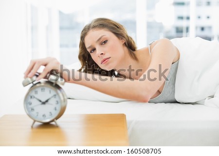 Annoyed beautiful woman turning off the alarm clock while lying in her bed and looking at camera