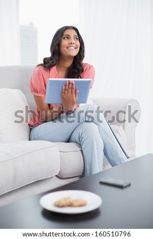 Happy cute brunette sitting on couch holding tablet in bright living room