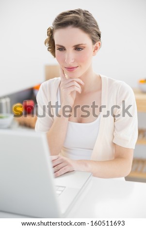 Attractive calm woman sitting thoughtful in front of her laptop in her kitchen