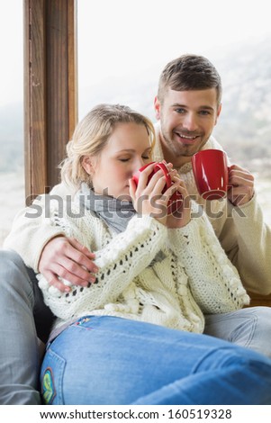 Loving young couple in winter clothing drinking coffee against cabin window