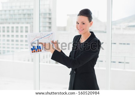 Portrait of a smiling elegant businesswoman pointing at graphs at bright office