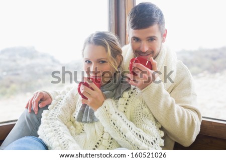 Portrait of a young couple in winter clothing drinking coffee against cabin window