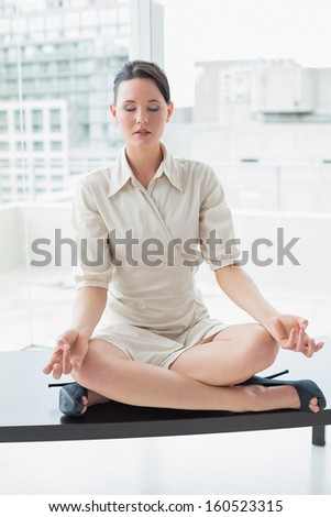 Full length of an elegant young businesswoman sitting in lotus position with eyes closed at bright office