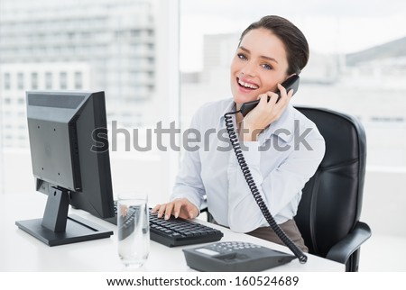 Portrait of a smiling elegant businesswoman using landline phone and computer in a bright office