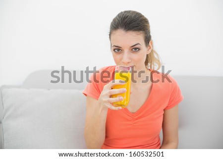 Gorgeous calm woman drinking a glass of orange juice looking at camera sitting on couch