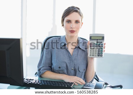 Calm businesswoman showing calculator sitting at her desk looking at camera