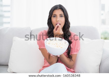 Frowning cute brunette sitting on couch holding popcorn bowl in bright living room