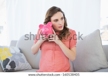 Lovely pregnant woman shaking a piggy bank sitting on couch in the living room
