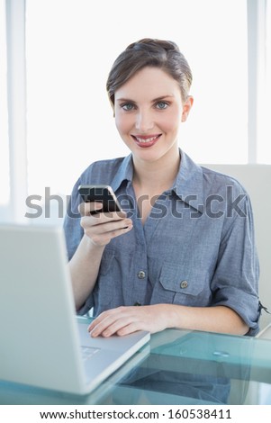 Portrait of pretty calm businesswoman holding her smartphone sitting at her desk in front of her laptop
