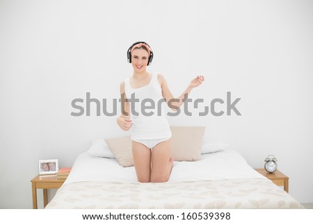 Woman playing air guitar while listening to music, kneeling on her bed and looking into the camera