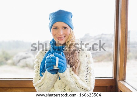Portrait of a cute young woman with coffee cup in warm clothing against cabin window