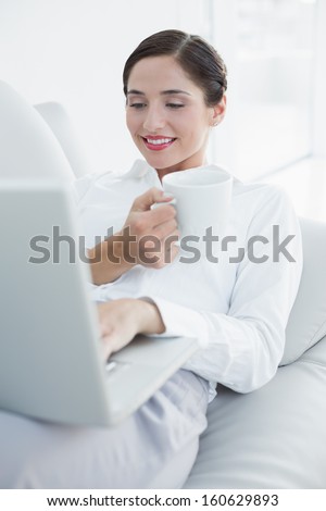 Smiling well dressed young woman with laptop and coffee cup on sofa at home