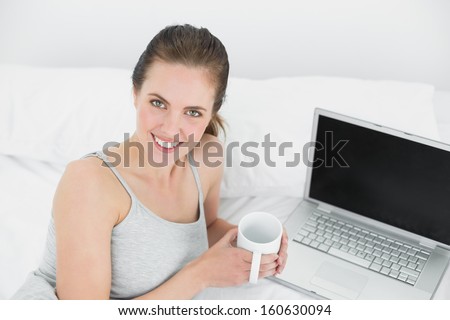 Portrait of a smiling casual young woman with laptop and coffee cup in bed at home