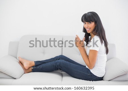 Gleeful young woman holding a cup sitting on couch smiling at camera