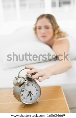 Pretty annoyed blonde lying in bed turning off alarm clock in bright bedroom