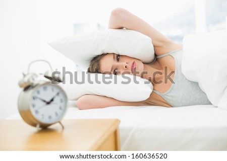Tired young woman lying in her bed covering her ears with pillow looking at the alarm clock