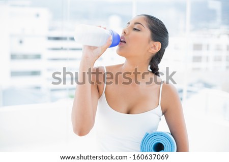 Toned brunette drinking from sports bottle and holding exercise mat in bright room