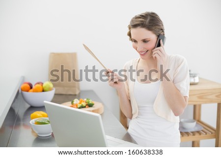 Content woman phoning with her smartphone in the kitchen looking at her laptop