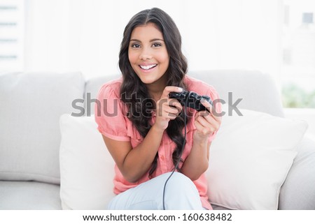 Happy cute brunette sitting on couch holding controller in bright living room