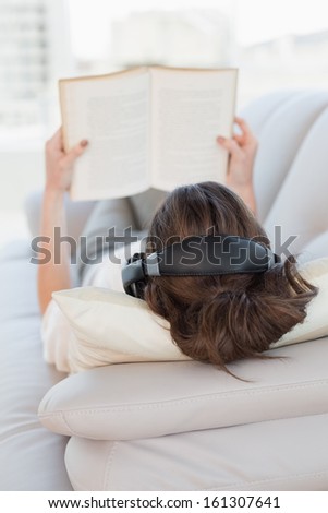 Close up of a brown haired casual woman enjoying music while reading a book on sofa in a bright house