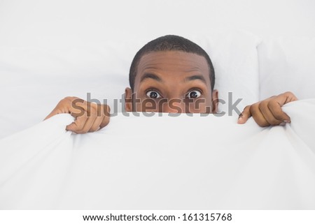 Close up portrait of a young shocked Afro man covering face with sheet in bed at home