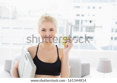 Portrait of a smiling fit young woman holding scale and apple in fitness studio
