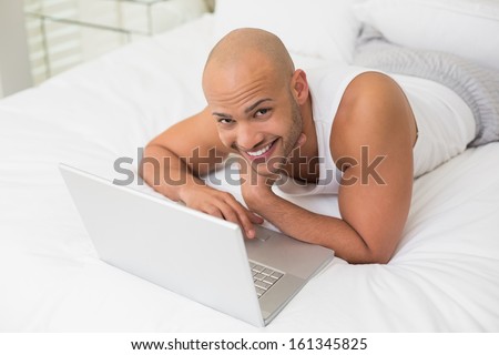 Portrait of a smiling casual bald young man using laptop in bed at home