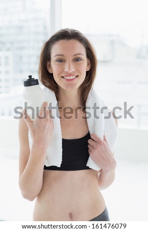 Portrait of a young woman with towel around neck holding water bottle in fitness studio