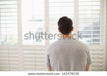 Rear view of a young man looking through window blinds at a bright room