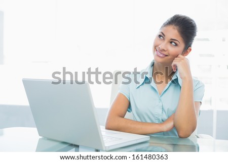 Thoughtful businesswoman looking up in front of laptop on desk in a bright office