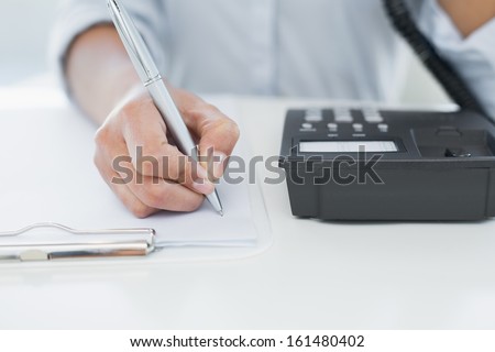 Close Up Mid Section Of A Businesswoman Using Telephone While Writing On Clipboard At Desk