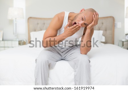 Young bald man sitting and yawning in bed at home