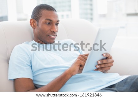 Side view of a casual young Afro man using digital tablet on sofa in a bright house