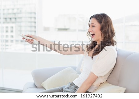 Side view of a cheerful young woman with remote control sitting on sofa at bright home