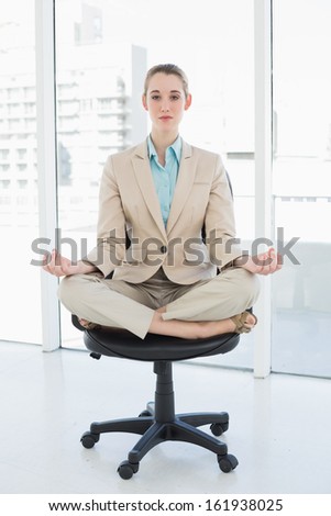 Focused chic businesswoman sitting in lotus position on her swivel chair looking at camera