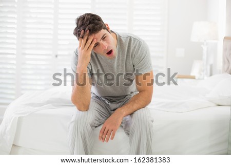Sleepy young man sitting and yawning in bed at home