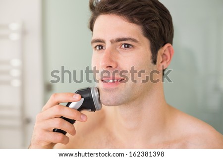 Smiling handsome young shirtless man shaving with electric razor