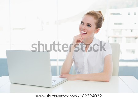 Thoughtful businesswoman looking up while using laptop on desk in a bright office