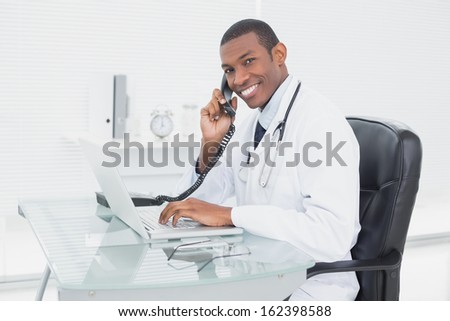 Side view portrait of a smiling male doctor using phone and laptop at medical office